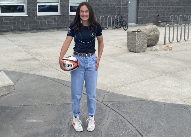 A female athlete wearing jeans and a T-shirt, holding a rugby ball in a casual setting in front of her high school