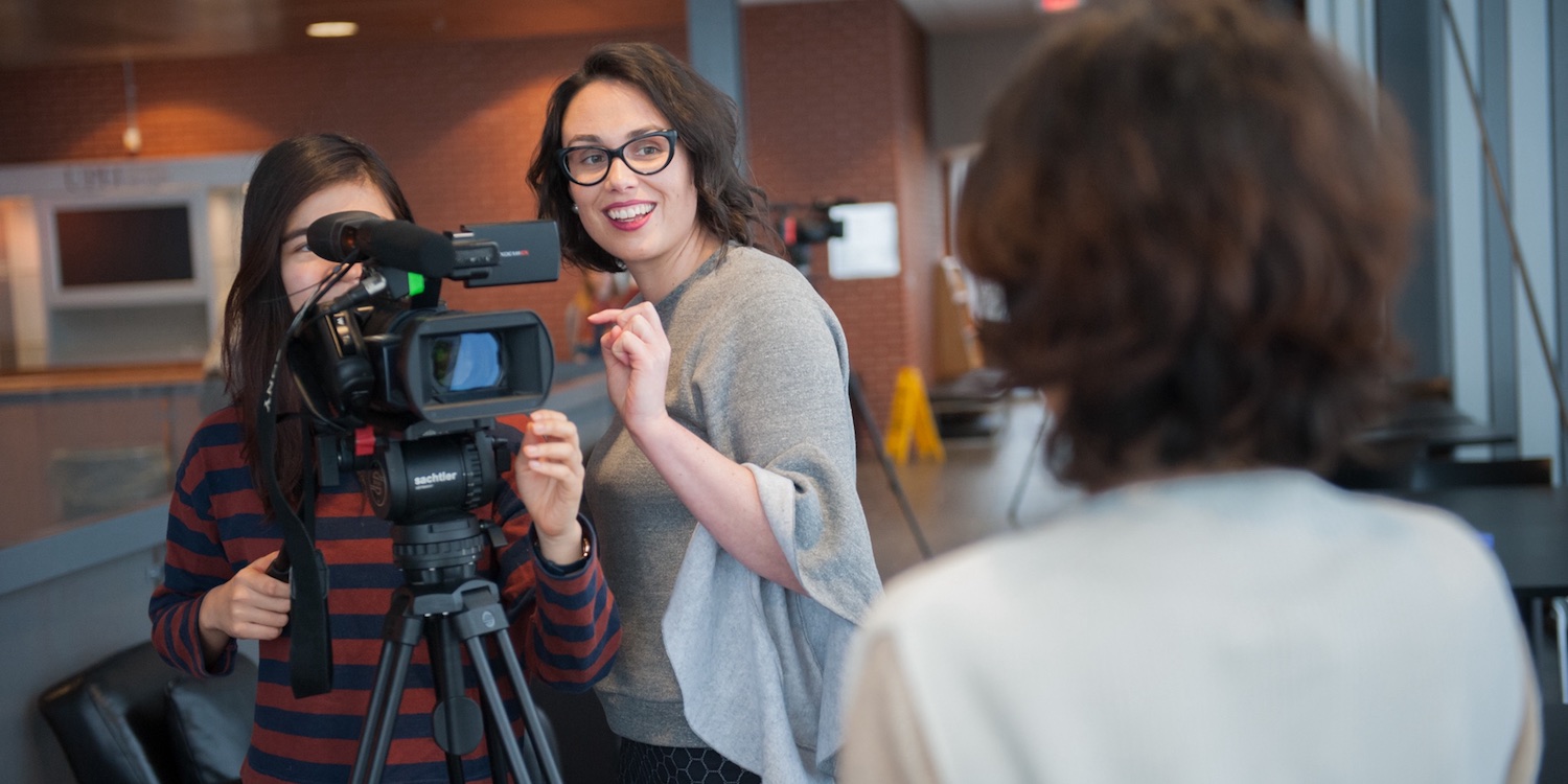two people behind a video camera with a female subject in the foreground