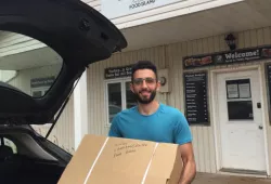 A smiling male student loads a large box into the back of a vehicle