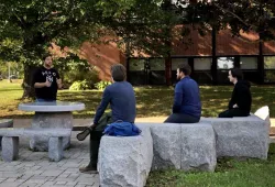 Three students sit on stone slabs in an outdoor setting as they listen to a fourth student instructing them