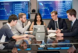A group of students have an animated discussion around a table with computers, with displays in the background of charts and graphs