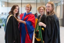 A mature woman in red and blue academic robe helps three younger woman as they put the finishing touches putting on their own academic robes