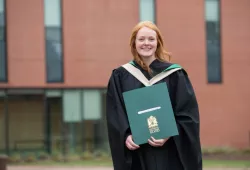 A smiling woman with red hair in graduation gown grips her bachelor's degree