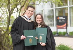 A smiling male and female wearing academic robes clutch embossed folders with the crest of UPEI on the cover