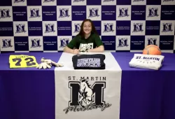 A female soccer player wearing a UPEI Panthers t-shirt sits at a table covered in logos and jerseys of her high school, St. Martins Secondary School