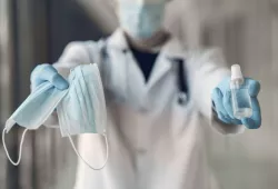 Image of person wearing lab coat, gloves, and a mask and holding PPE and sanitizer