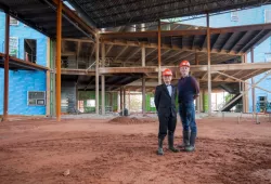 Two men, one wearing a tuxedo, another in more casual clothes, pose in a construction site with hard hats on 