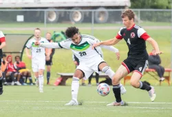 A male soccer player in green and white soccer gear attempts to steal a ball away from a defending player in a black and red uniform