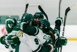 Several female hockey players in green and white Panther hockey gear celebrate a goal with an on-ice embrace