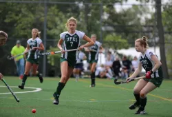 A female athlete in a green and white panthers uniform chases a ball down a grass field with a hockey stick