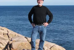 An older man in jeans and a long black t-shirt stands on the rocks beside the sea
