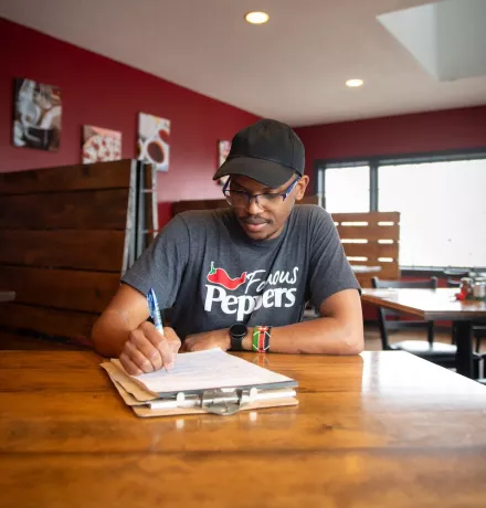 a smiling man sitting at a restaurant table writing on a clipboard