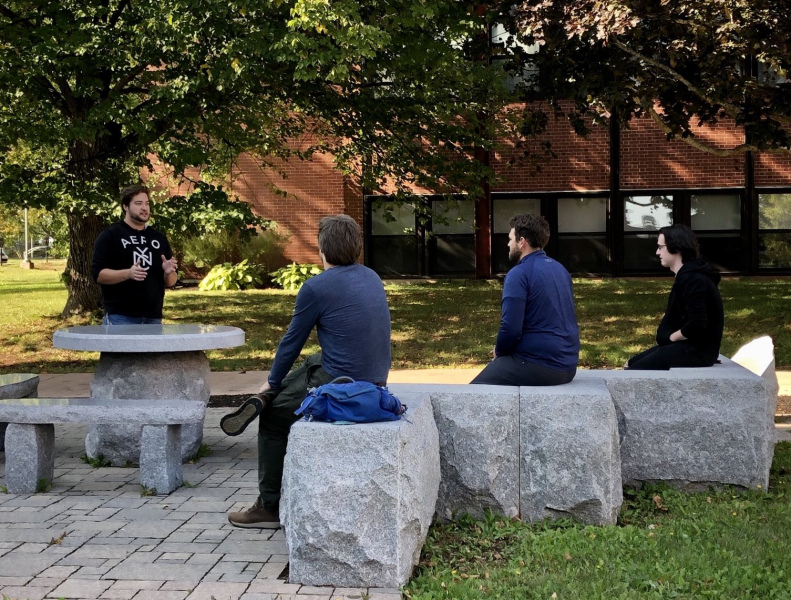 BEd students Gabriel Young, Colin Jeffrey, Connor Myers, and Spencer Stetson participate in an outdoor class, practicing their presentation skills. (Photo by Dr. Carolyn Thorne) Three students sit on stone slabs in an outdoor setting as they listen to a fourth student instructing them