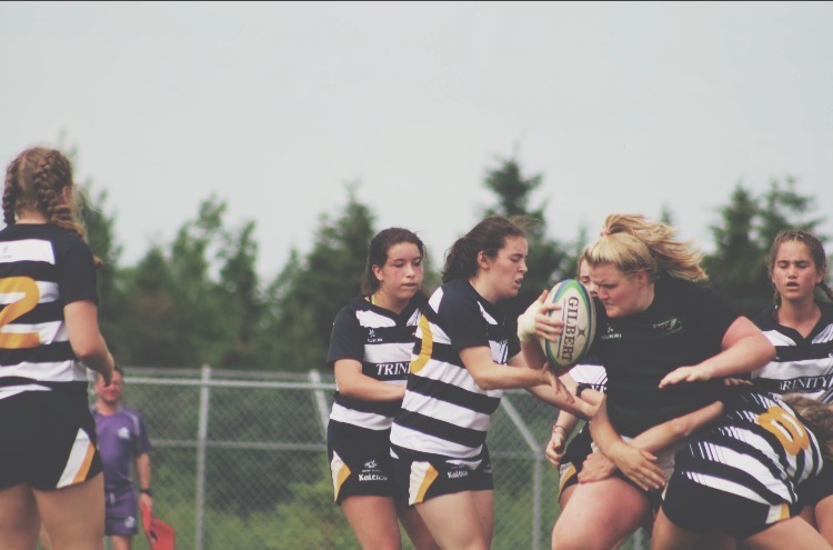 Emily-Anne MacQuarrie holds possession of the ball through a wall of defenders A woman in a black uniform holding a rugby ball charges through a group of women wearing striped uniforms