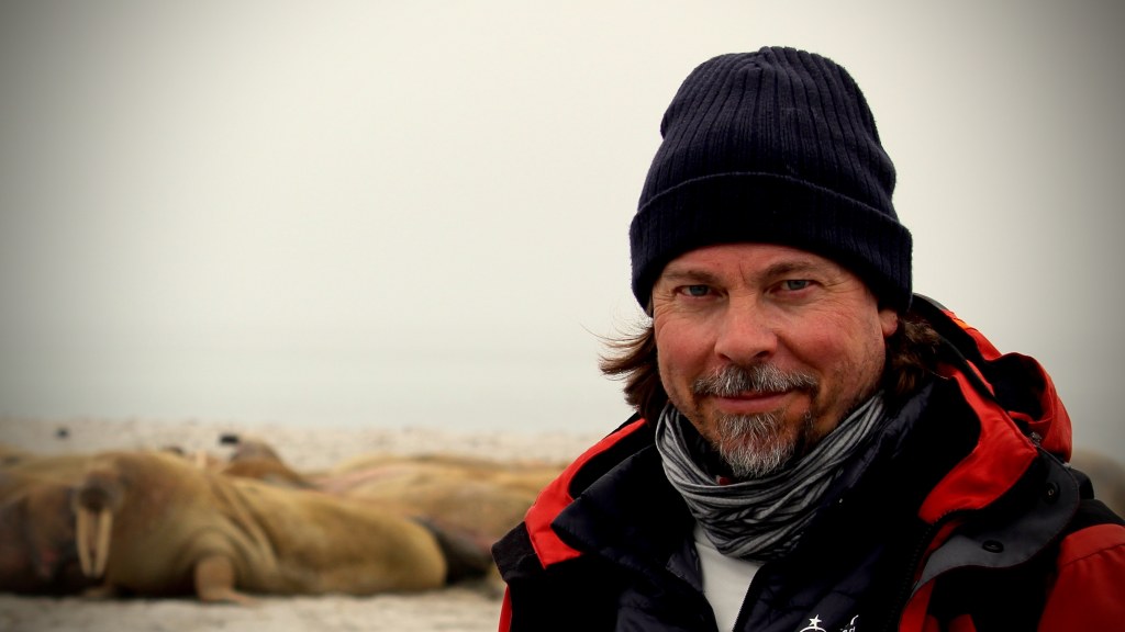 A medium close-up shot of a bearded man in winter hat and jacket in front of a chilly landscape