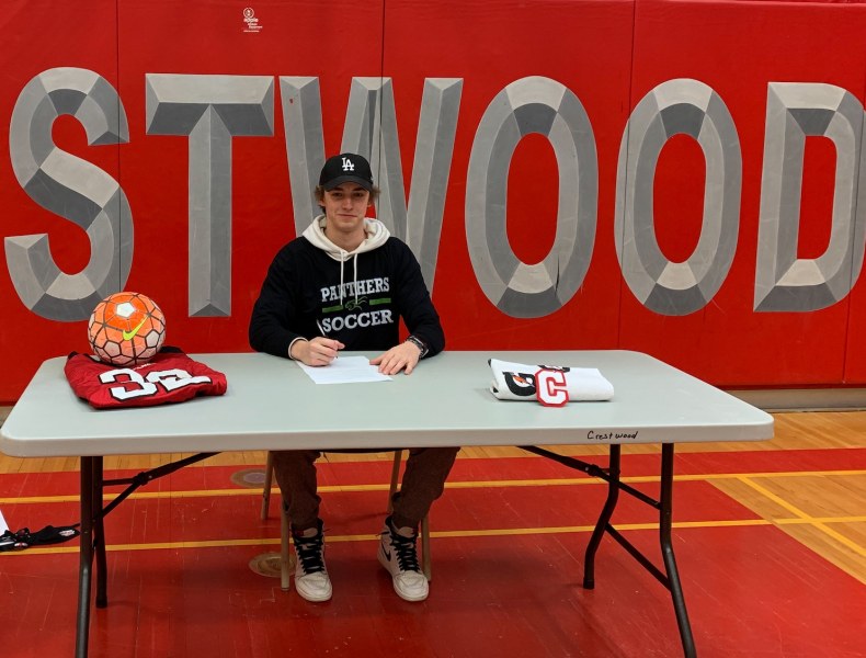 A smiling teenaged male sits at a table with the large logo fo CRESTWOOD secondary school behind him