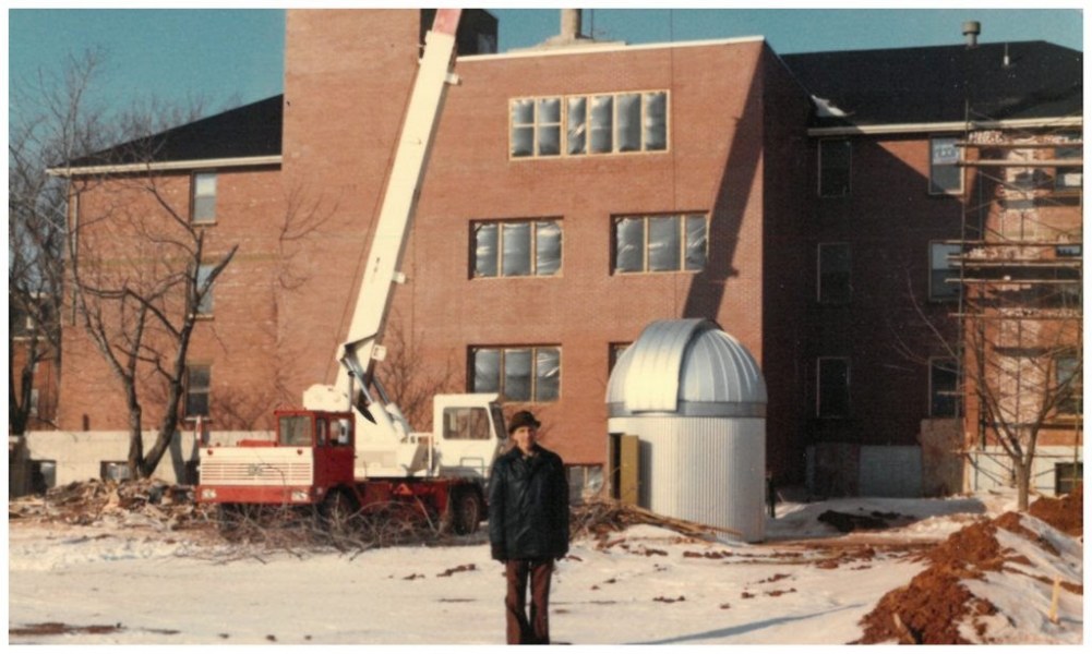 An aged photograph of a man standing next to a silver dome on the ground next to a brick building