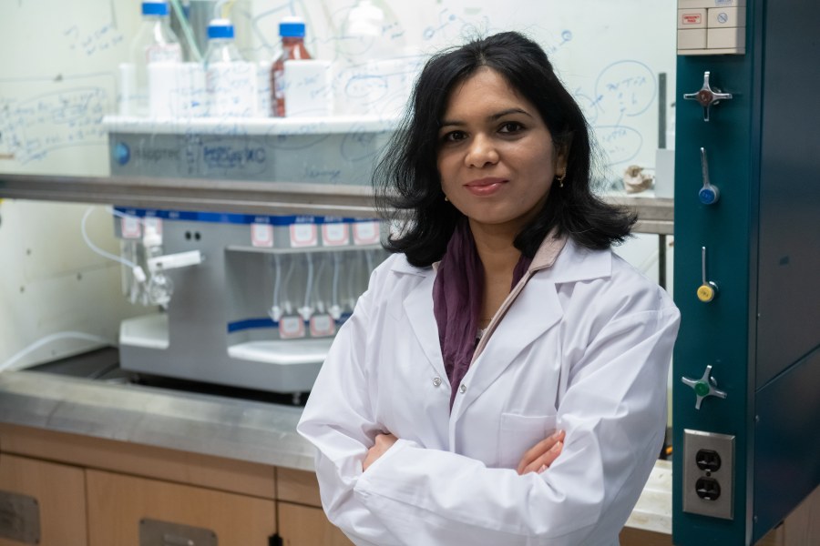 A female scientist in a white lab coat stands with her arms crossed in front of a laboratory environment in which complicated-looking scientific formulae are written out on a glass wall