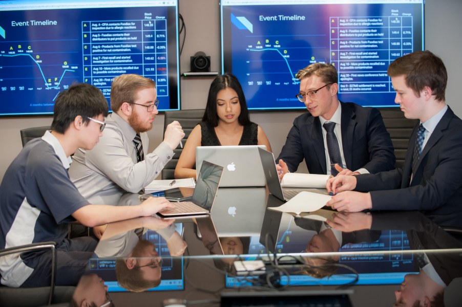 A group of students have an animated discussion around a table with computers, with displays in the background of charts and graphs