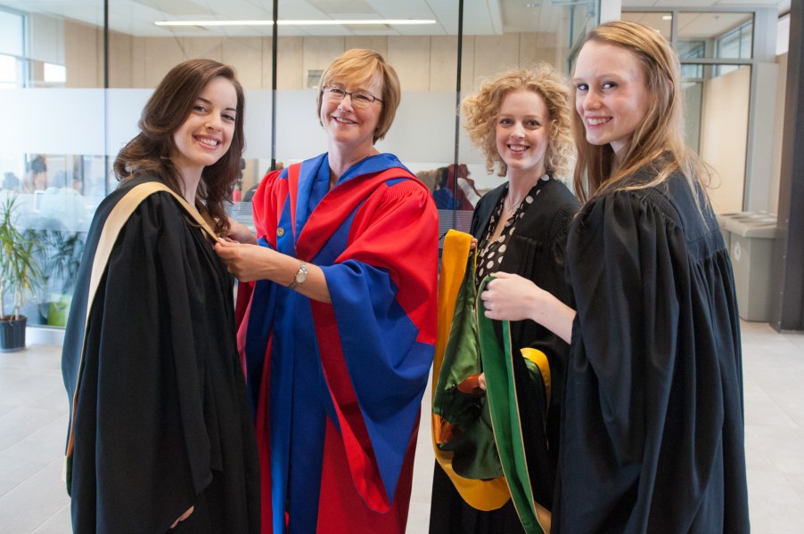 A mature woman in red and blue academic robe helps three younger woman as they put the finishing touches putting on their own academic robes