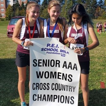 Three young females wearing matching running gear from Colonel Gray High School stand smiling with medals around their necks, holding a banner reading: SENIOR AAA WOMEN'S CROSS COUNTRY CHAMPIONS