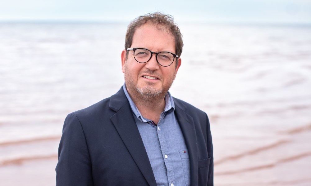Dr. Adam Fenech A smiling man in a jacket and unbuttoned shirt. Photo taken at the beach.