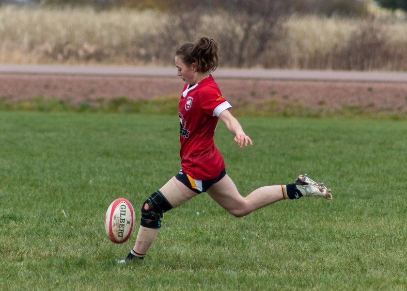 A female athlete extends her leg to kick a rugby ball on a grassy pitch