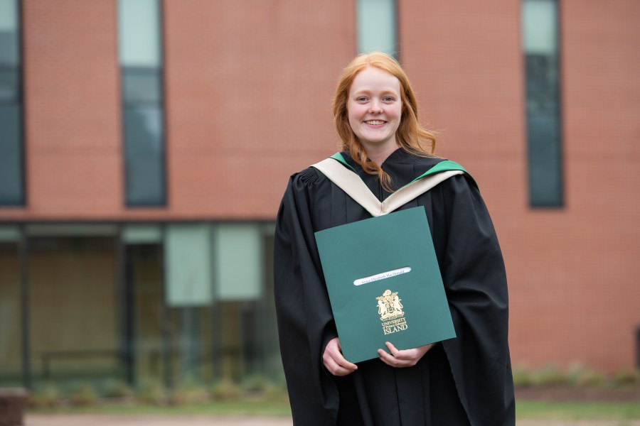 A smiling woman with red hair in graduation gown grips her bachelor's degree