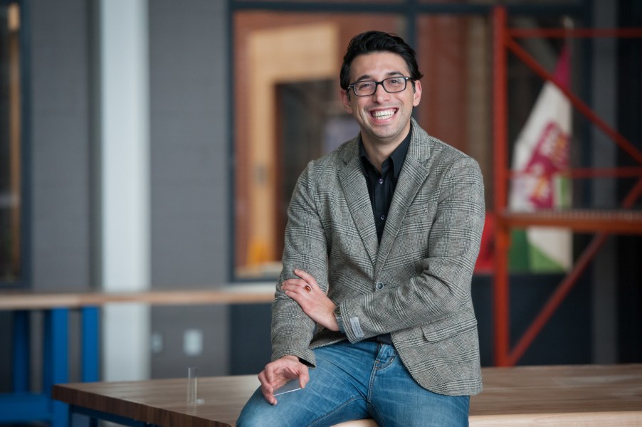 A smiling man in a jacket and unbuttoned shirt. He sits casually on the corner of a workbench in an engineering lab.