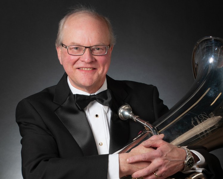 A smiling man in a black tuxedo cradles a tuba in his arms