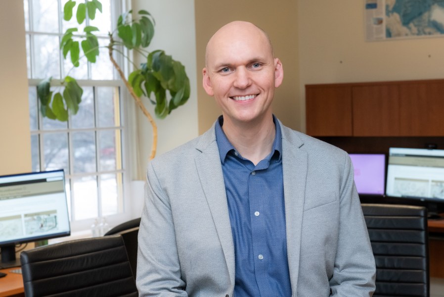 A smiling man in a jacket and unbuttoned shirt. He is sitting in an office with natural light and office plants