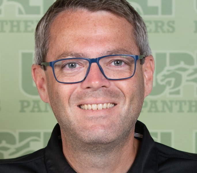 A smiling headshot of a man in glasses imposed over a a stylized UPEI Panthers background