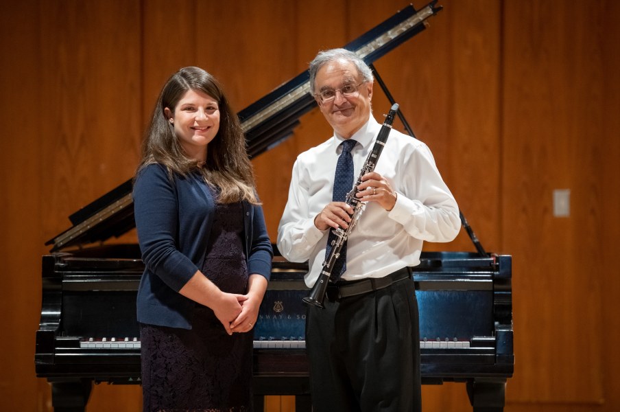 A smartly dressed woman with her hands crossed beside a man in a tie holding a clarinet. They both stand in a concert hall in front of a grand piano.