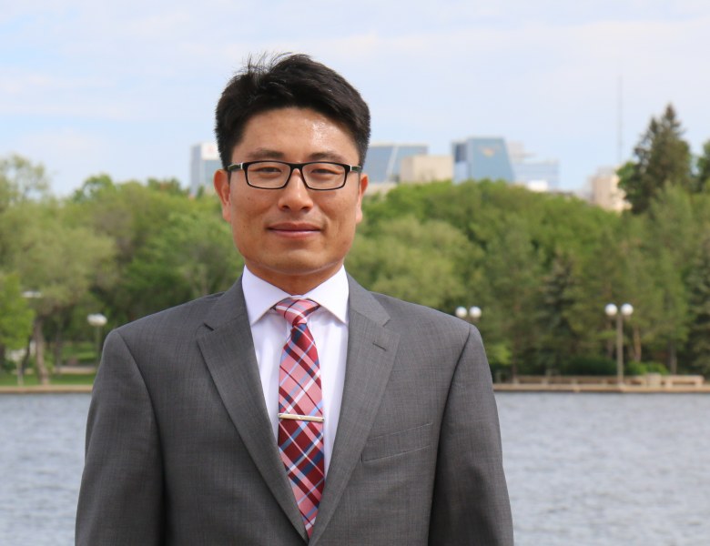 A professional-looking man n a grey suit stands in front of a river with trees and buildings on the opposite shore