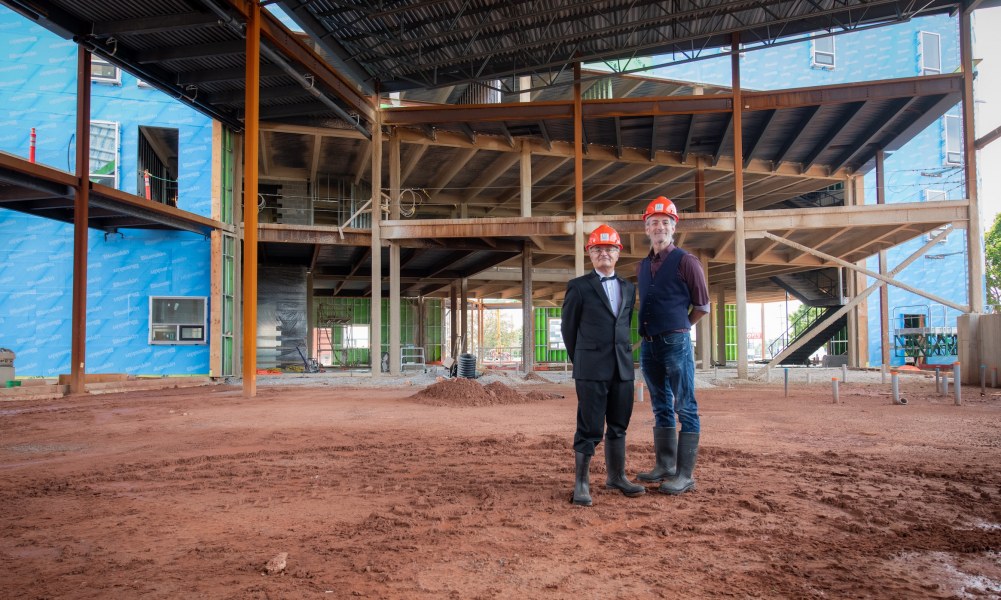 Two men, one wearing a tuxedo, another in more casual clothes, pose in a construction site with hard hats on 