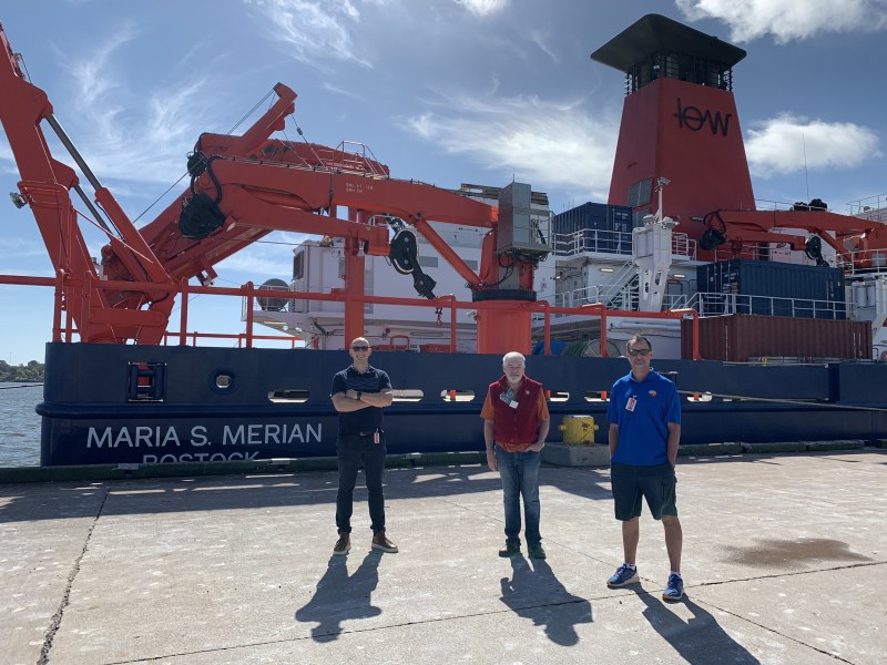 Three smiling men stand on a wharf next to a giant ship festooned with cranes and other research material