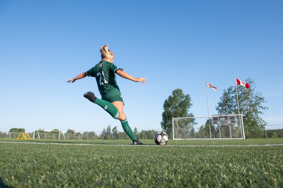 A female soccer player in a green Panthers soccer uniform winds up with her right foot to take a shot on net
