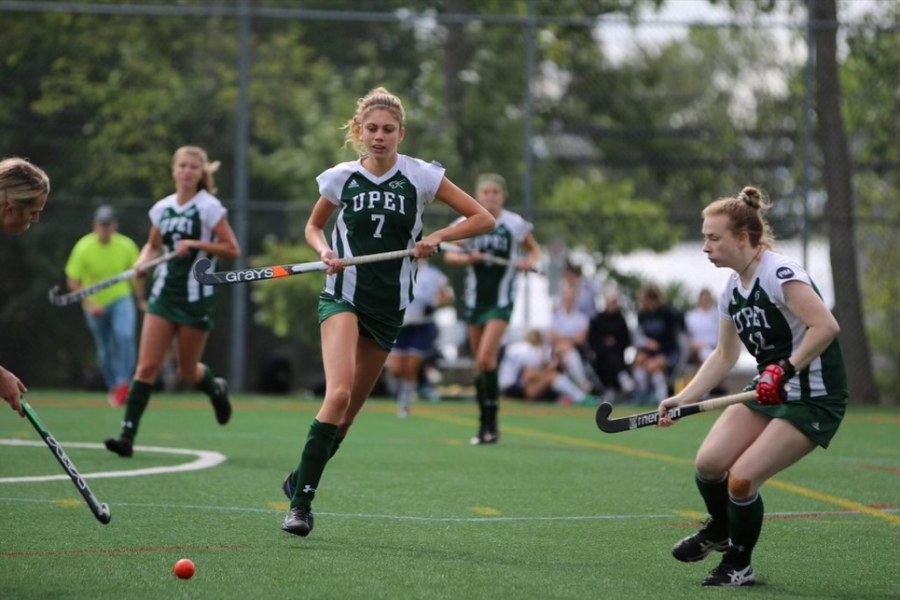 A female athlete in a green and white panthers uniform chases a ball down a grass field with a hockey stick