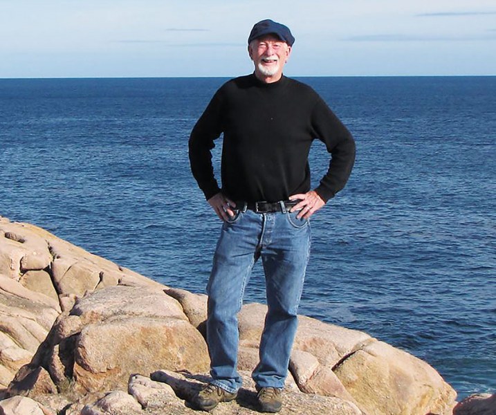An older man in jeans and a long black t-shirt stands on the rocks beside the sea