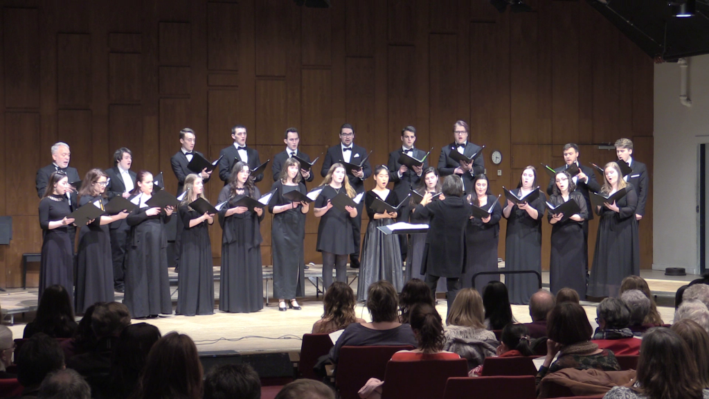 A small chamber choir on stage wearing black concert robes
