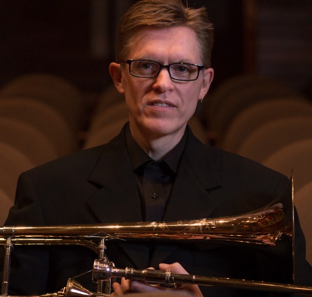 A man in dark-framed glasses and black performing clothes cradles a trombone on his lap