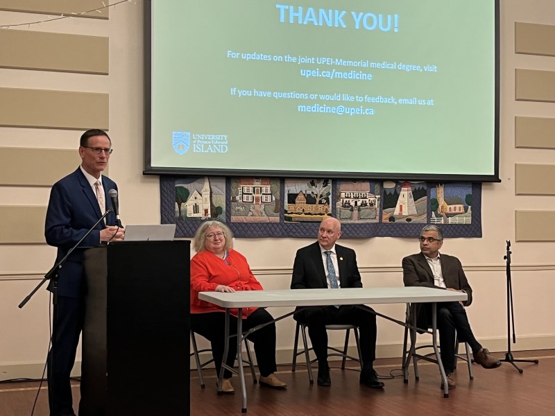 photo of man speaking at podium with three people sitting at an adjacent table