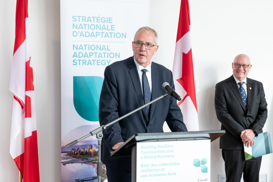 President Greg Keefe looks on as Hon. Bill Blair, Minister of Emergency Preparedness, announces Canada's National Adaptation Strategy in St. Peter's Bay man speaking at podium flanked by two Canadian flags with man looking on