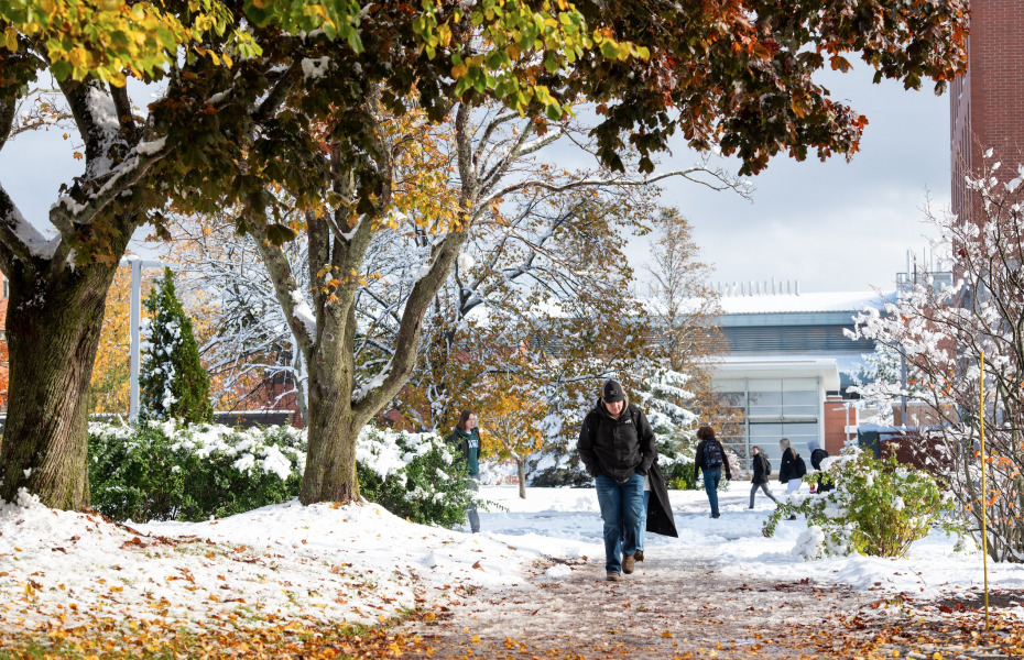 photo of man wearing ball cap walking on leaf and snow covered walkway on campus