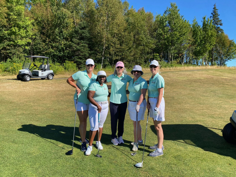 photo of five women wearing green shirts, standing on a golf course