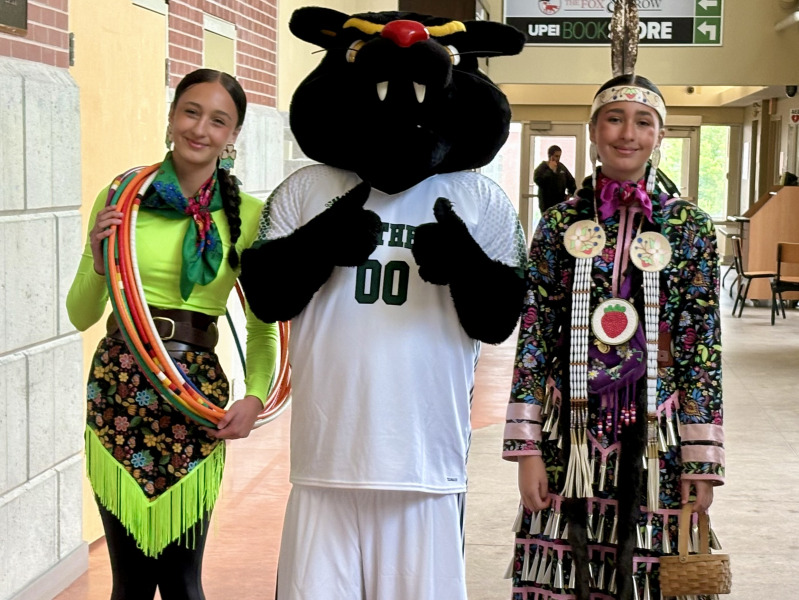 Two young women wearing Mi'kmaw regalia meet up with the UPEI mascot