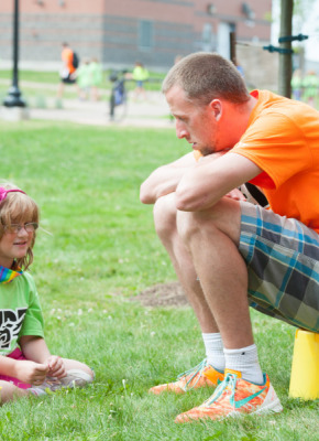A young girl sits on the grass speaking with an adult