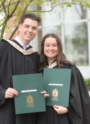 A smiling male and female wearing academic robes clutch embossed folders with the crest of UPEI on the cover