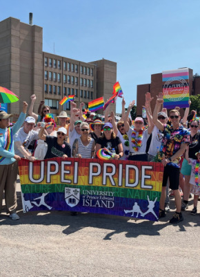 Members of the UPEI community at the 2024 PEI Pride Parade Photo of large group of people holding a banner and wearing clothing in bright rainbow colours