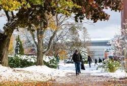 photo of man wearing ball cap walking on leaf and snow covered walkway on campus